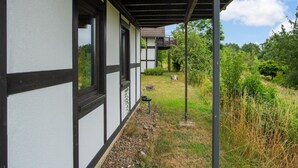 Una casa moderna con un porche cubierto, ventanas amplias y un jardín con terraza de madera y follaje.