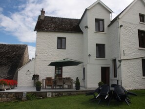 Un edificio blanco de dos pisos con base de piedra, techo de tejas y un pequeño patio con muebles de exterior y una sombrilla verde.