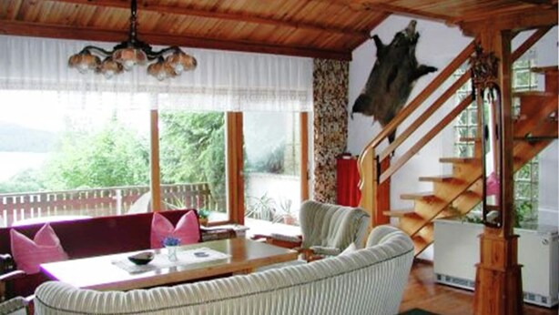 Ein Zimmer mit Holzbalkendecke, einem Esstisch mit Stühlen, einer Couch und einer Wendeltreppe, mit Blick auf eine grüne Landschaft.