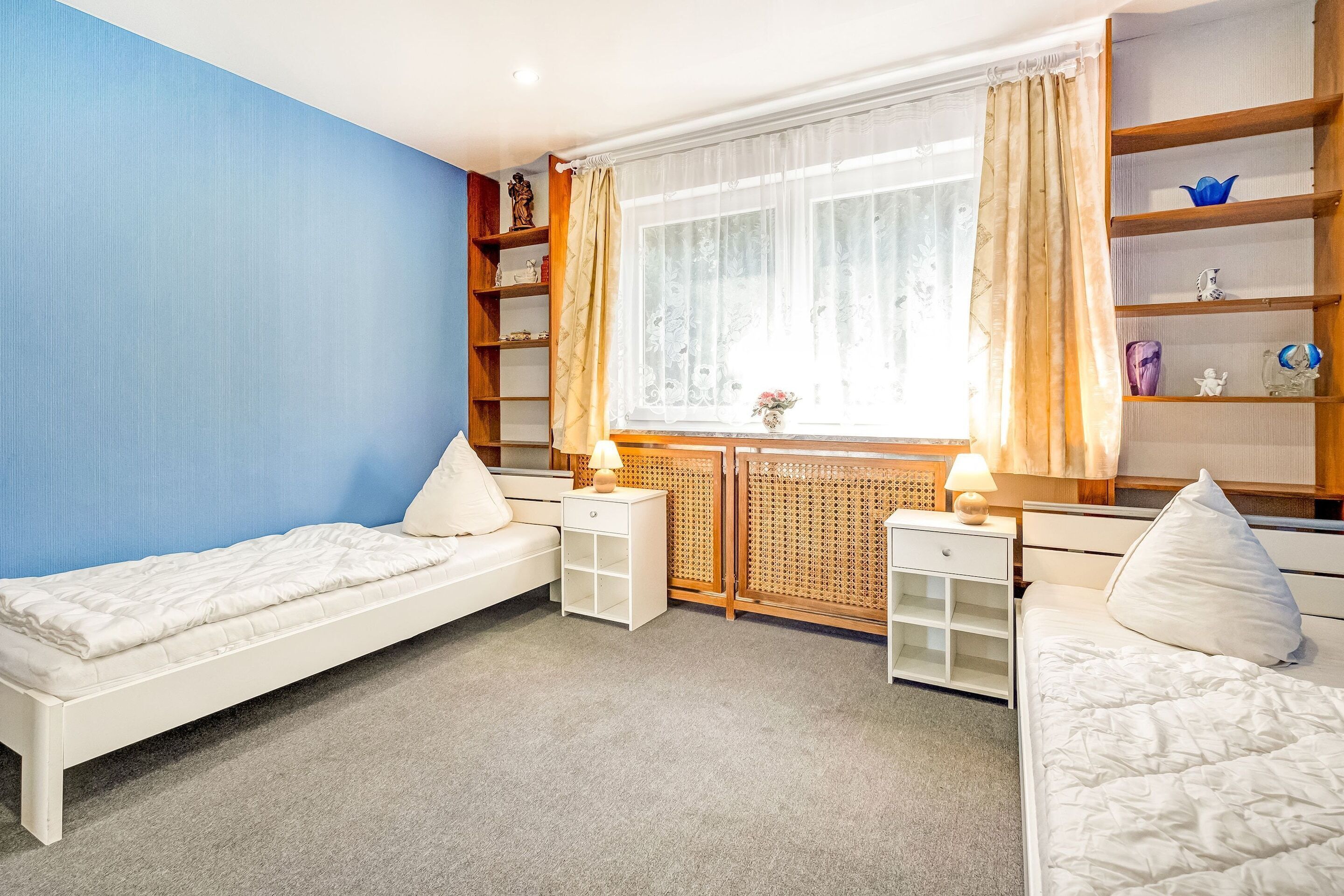 Ein Schlafzimmer mit zwei Einzelbetten, einem Fenster mit Vorhängen, einem Heizkörper und einem Bücherregal.