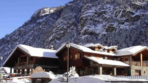 A wooden lodge with a snow-covered roof, situated in a mountainous area with a clear sky.