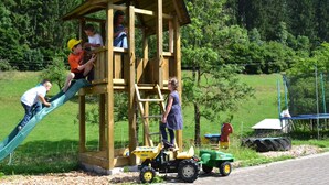 Una estructura de juego de madera con tobogán y escalera de cuerda, rodeada de follaje y un sendero pavimentado.