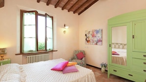 Een slaapkamer met een groot bed, een groene garderobe, een stoel met een bloemenkussen, een radiator en een raam met gordijnen.