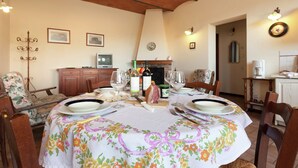 Een eetgedeelte met een ronde tafel gedekt voor vier personen, omringd door houten stoelen. Achtergrond is een open haard, en de kamer heeft een rustiek plafond van baksteen.