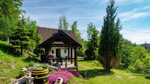 A wooden gazebo with a tiled roof, surrounded by a lush garden with purple flowers and a walking path.