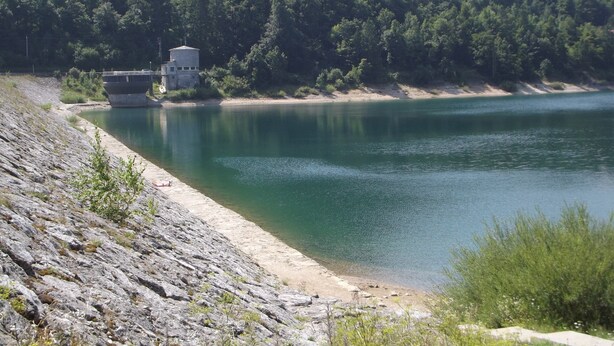 A lake with a concrete structure on the left, surrounded by dense forest and a rocky shoreline.