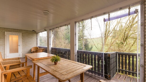 A wooden deck with a table and chairs, a potted plant, and a view of trees through the railing.