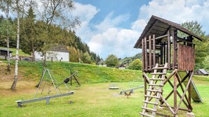 A rustic wooden structure with a ladder, a seesaw, and a swing set in a grassy area with trees and houses in the background. - Splendid Holiday Home in Untervalme near Ski Area (Bestwig)