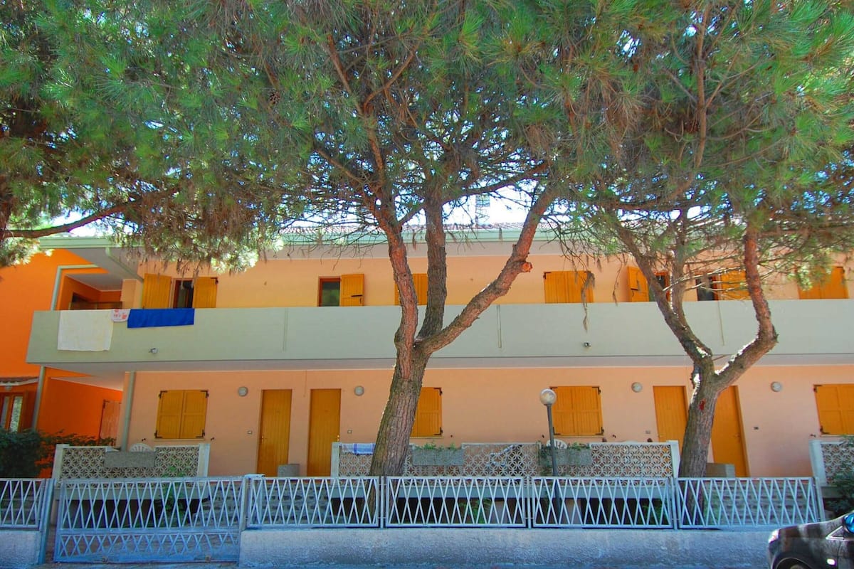 A two-story building with a balcony on the upper floor, a white fence in front, and trees in the background.