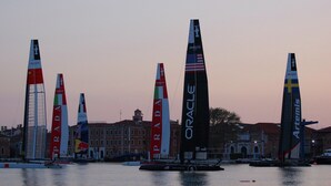 En hamn med många segelbåtar, varav några har väl synliga varumärkeslogotyper, till exempel Prada och Oracle.