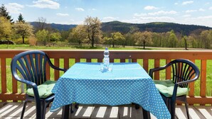 A table with a blue polka dot tablecloth, two chairs, and a bottle of water on a wooden deck with a view of a forest and mountains. - Gemutliches Ferienhaus in Sebnitz (Sebnitz)