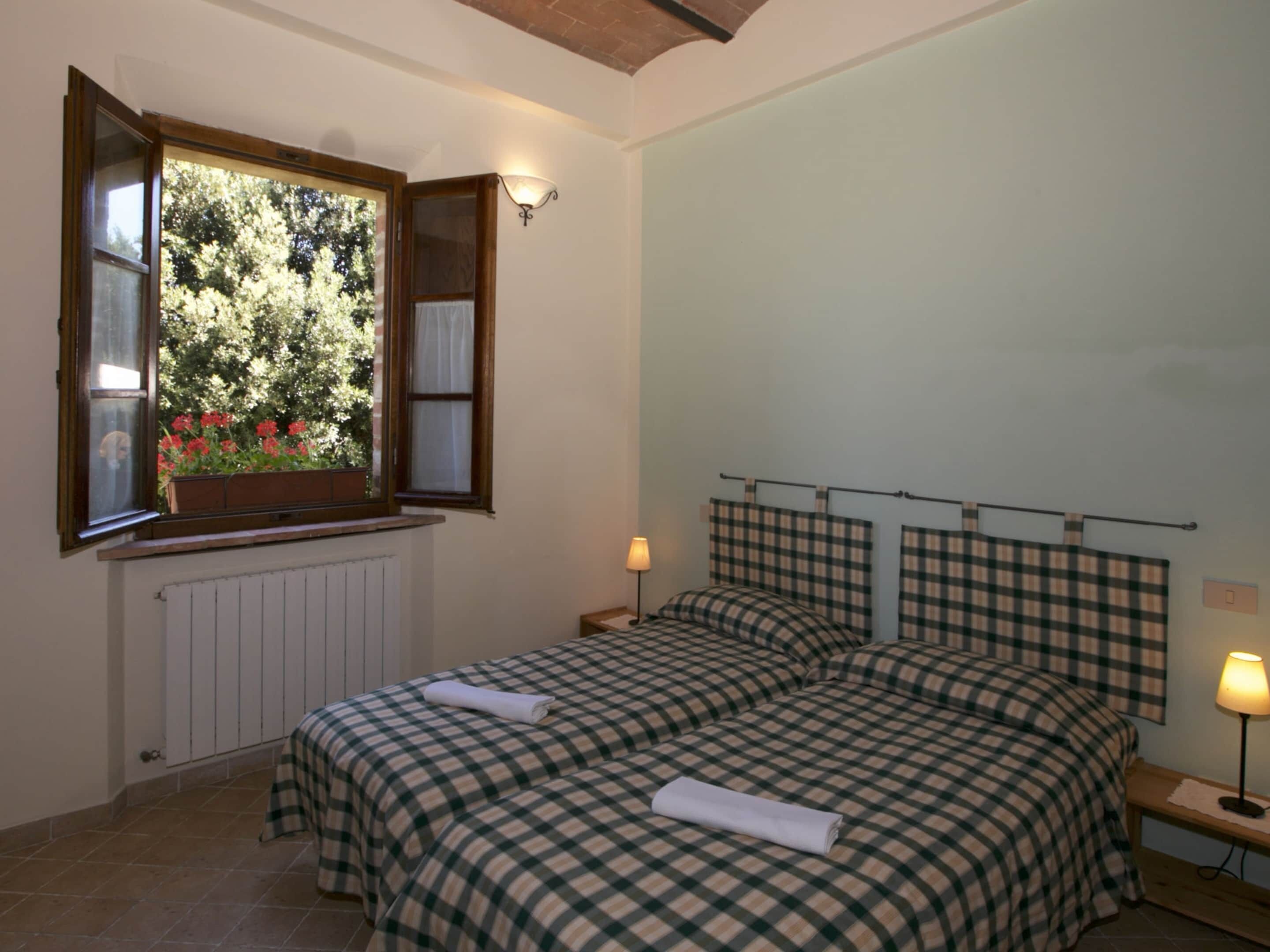 Una camera da letto con due letti, una finestra vista alberi e fiori, e un termosifone.
