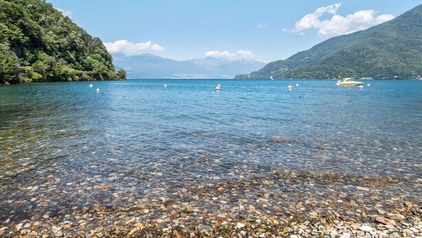 A clear lake with boats, pebbles, and mountains in the background. - Belvilla by OYO Studio Garden Uno Bis (Cannobio)