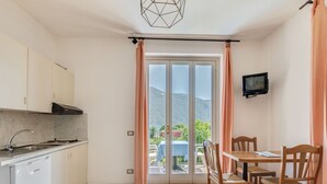 A compact kitchen with a built-in stove, a sink, and cabinets. There is a dining area with a table and chairs, and a TV mounted on the wall. A sliding glass door leads to a balcony with a view of the mountains. - Belvilla by OYO Studio Garden Uno Bis (Cannobio)