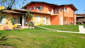 Un bâtiment à deux étages avec une façade rouge, un petit jardin devant avec une pelouse et un chemin menant à l’entrée.