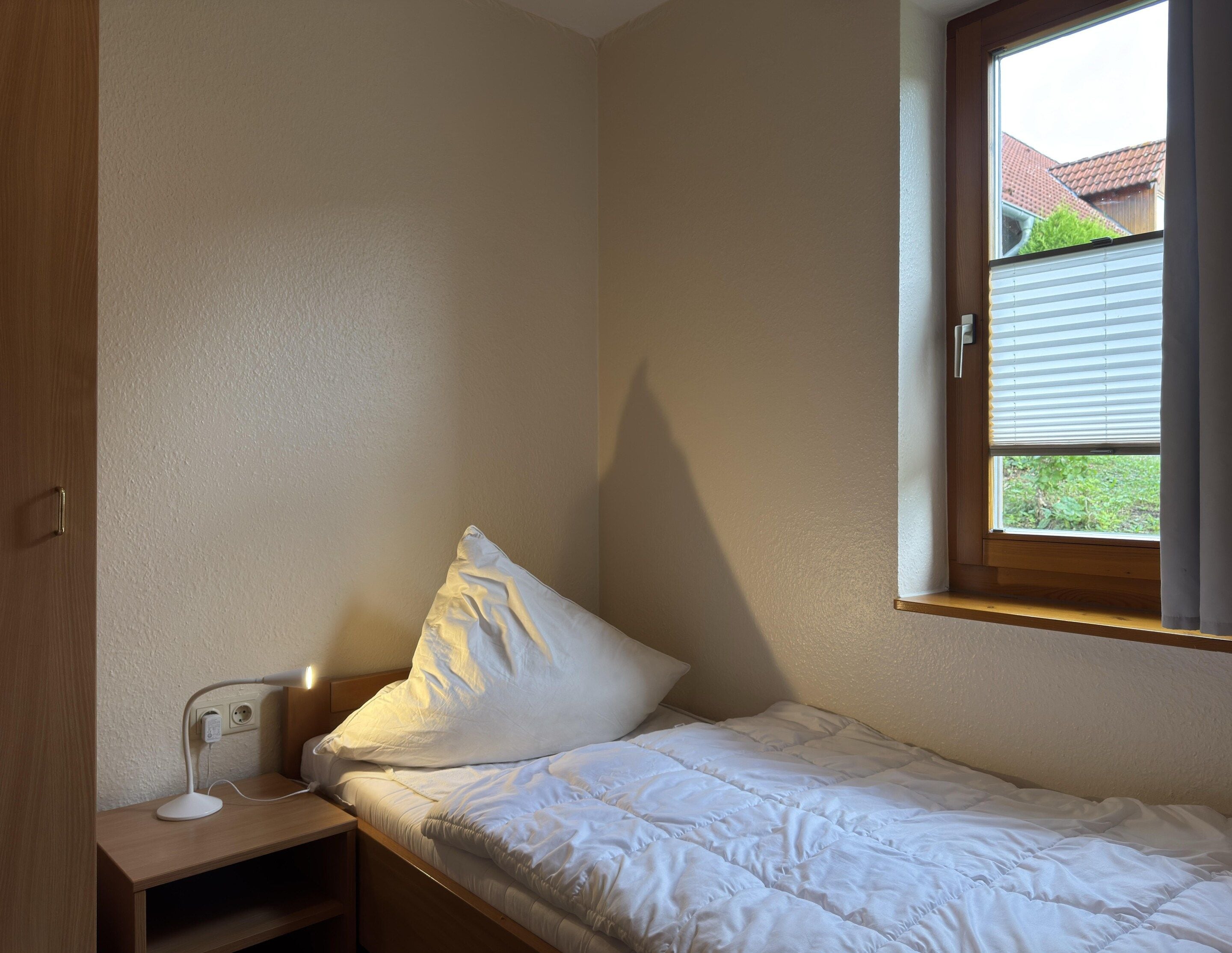 A small, single-bed room with a wooden headboard, a bedside table with a lamp, and a window with blinds.
