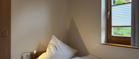 A small, single-bed room with a wooden headboard, a bedside table with a lamp, and a window with blinds.