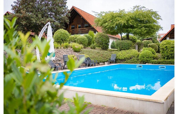 A swimming pool with lounge chairs, surrounded by well-maintained bushes and trees, with a wooden building in the background.