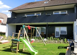 Ein Wohnhaus mit dunkler Fassade, einer Veranda und einem Hinterhof mit einer Holz-Schaukel und einer grünen Rutsche.