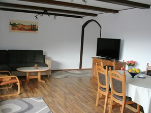 Ein Wohnzimmer mit einer Couch, einem runden Tisch, einem Fernseher und einem Esstisch mit Stühlen.