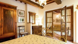 A bedroom with a bed, wooden wardrobe, and a chair. There is a door leading to another room with a sofa and curtains. - Holiday Home in Cortona (Cortona)