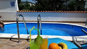 Escena junto a la piscina con piscina, dos bebidas con pajitas y rodajas de lima, un cuenco de fruta y una vela.