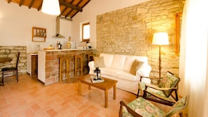 A cozy living area with a stone wall, a white sofa, a wooden coffee table, and a kitchen with a stone countertop and wooden stools. - Agriturismo Poggio di Montepescoli - Il Ciliegio (Pelago)