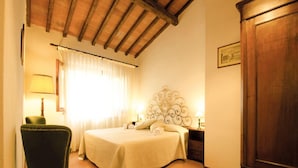 A bedroom with a bed, a chair, a nightstand, a lamp, a wardrobe, and a window with curtains. - Agriturismo Poggio di Montepescoli - Il Ciliegio (Pelago)