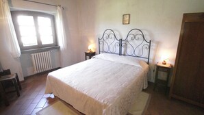 A bedroom with a bed, two bedside tables with lamps, a radiator, a window with curtains, and a door.