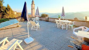An outdoor terrace with white plastic chairs and tables, a blue umbrella, and a view of a clock tower and mountains. - Holiday Home in Sesta Godano With BBQ (Sesta Godano)