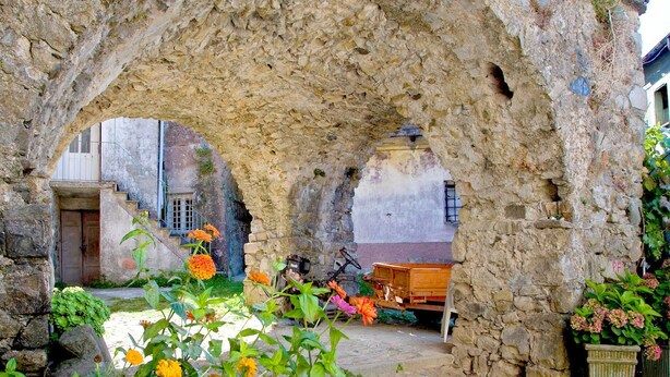 An ancient stone archway with a cobblestone courtyard, surrounded by flowering plants and a rustic wooden bench. - Holiday Home in Sesta Godano With BBQ (Sesta Godano)