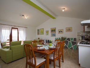 A dining area with a wooden table set for four, surrounded by wooden chairs. There is a green sofa and a dining table with a green and white geometric backsplash. The room has white walls and a sloped ceiling with recessed lighting. - Luxurious Apartment in Sajini Croatia (Barban)