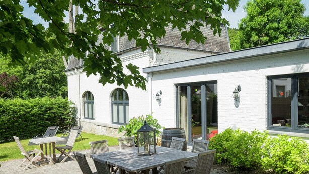 A patio with a wooden table and chairs, surrounded by greenery and a white building with arched windows. - Namur Mansion by the Meus (Hastiere)