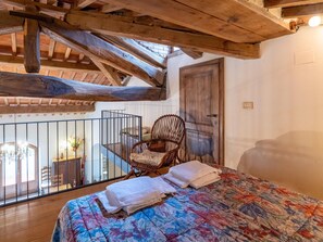 A bedroom with a wooden ceiling, a bed with a floral duvet, a wicker chair, and a view of a balcony with a railing. - Belvilla by OYO Casa Mirolago (Caprese Michelangelo)