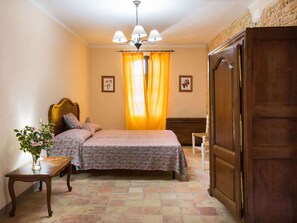A bedroom with a bed, a nightstand, a wooden wardrobe, a chandelier, and a window with curtains. - Belvilla by OYO Casa de Poi (Salsomaggiore Terme)