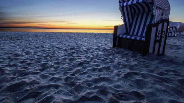 Ein Liegestuhl mit gestreifter Auflage und einer Nummer auf einem sandigen Strand, im Hintergrund ein Sonnenuntergang.