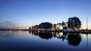 Ein Yachthafen im Dämmerlicht, das ruhige Wasser spiegelt Boote und Bäume wider.