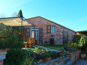 Une maison de campagne en briques, dotée d’une porte en bois et de fenêtres à volets, entourée d’un jardin bien entretenu où sont disposées diverses plantes en pot.