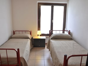 A small, clean room with two single beds, a nightstand with a lamp, and a window with a view of the outside.