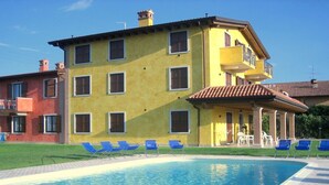 A two-story building with a swimming pool, blue lounge chairs, and a covered patio area. - Belvilla by OYO Casa Gialla A Due (Lazise)