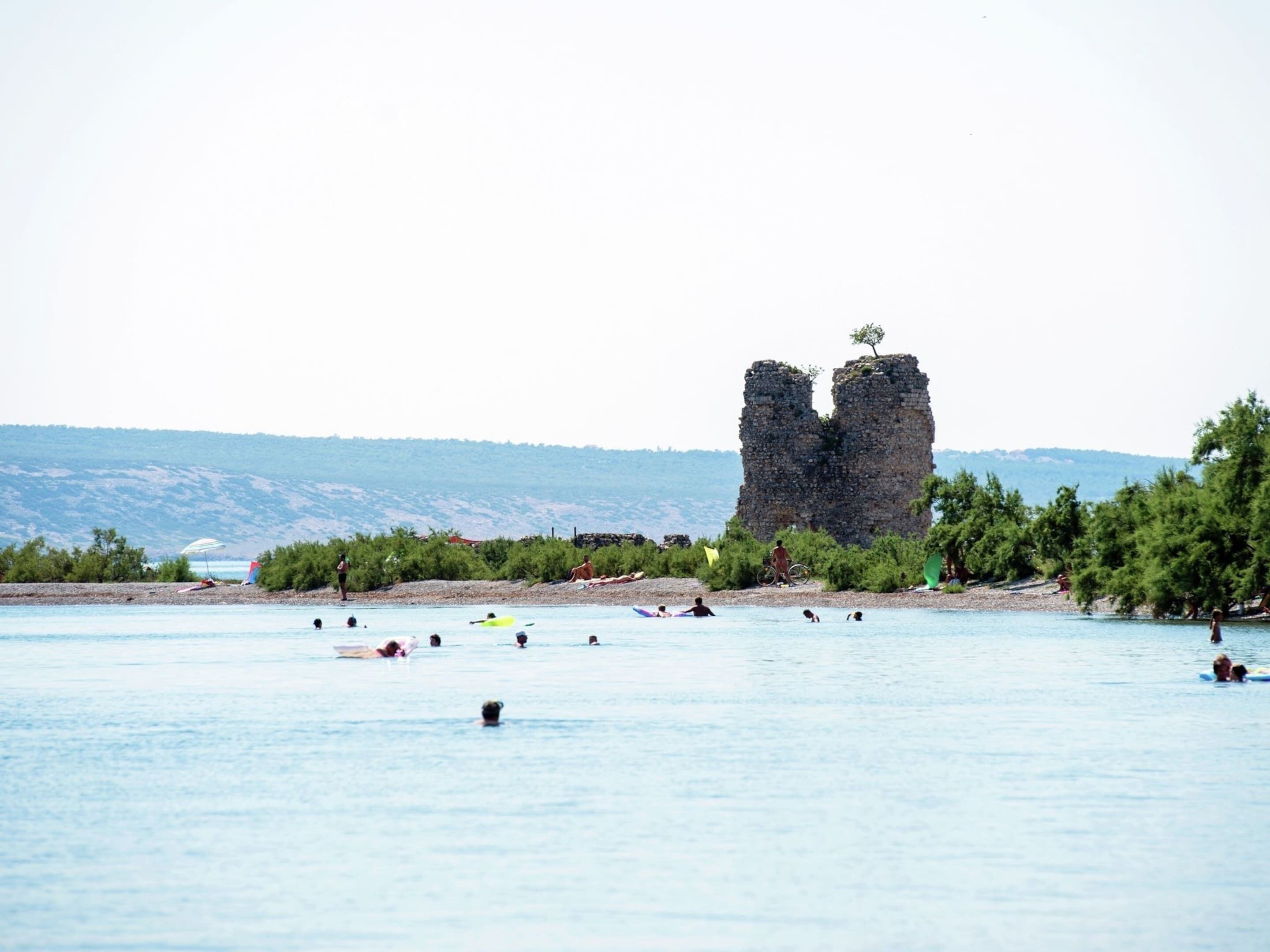 A beach with a large rock formation in the distance, surrounded by greenery and a calm sea.