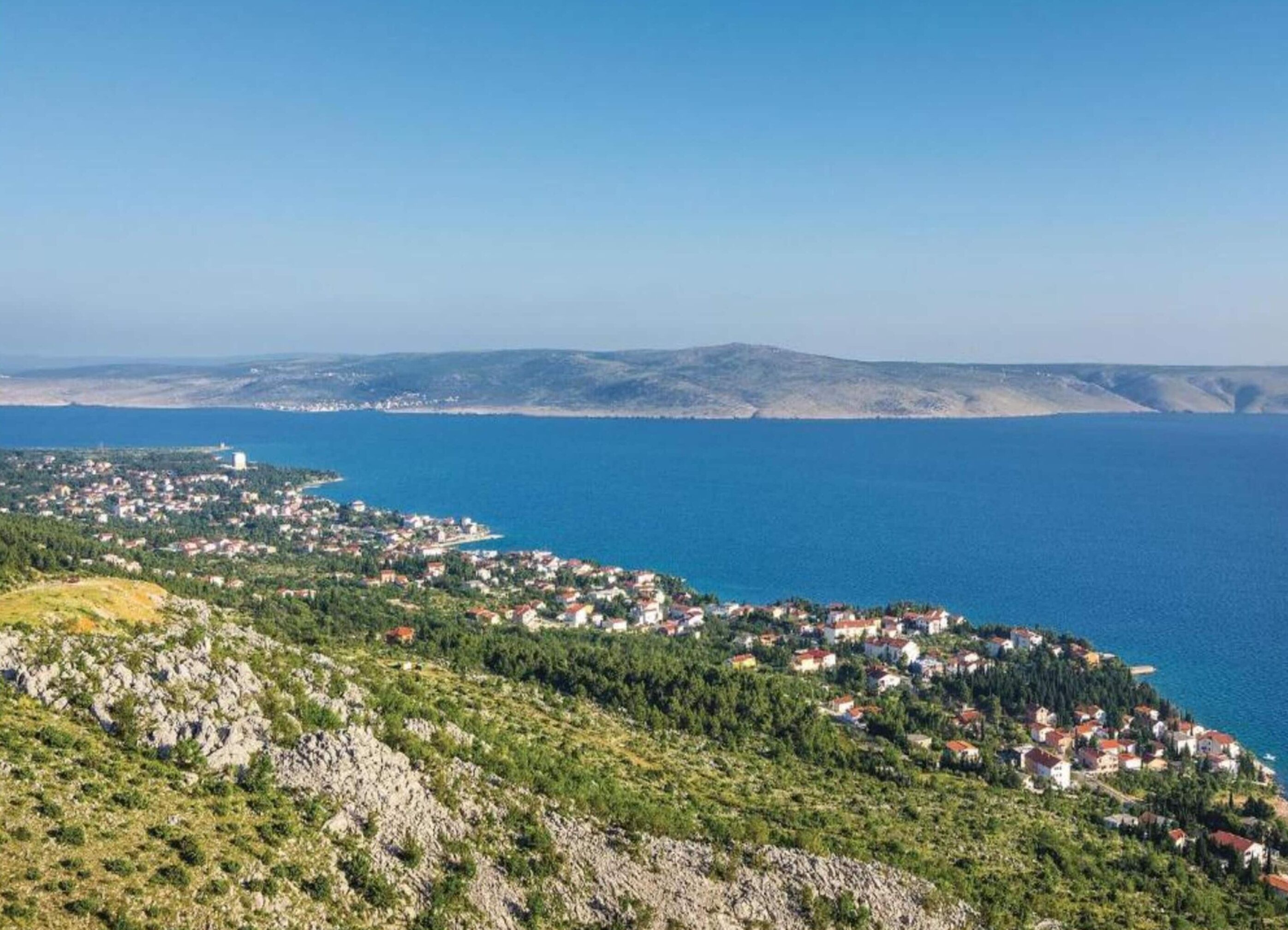 Une ville côtière où les bâtiments sont regroupés en grand nombre le long du littoral, avec un imposant château d’eau et un paysage vallonné en arrière-plan.
