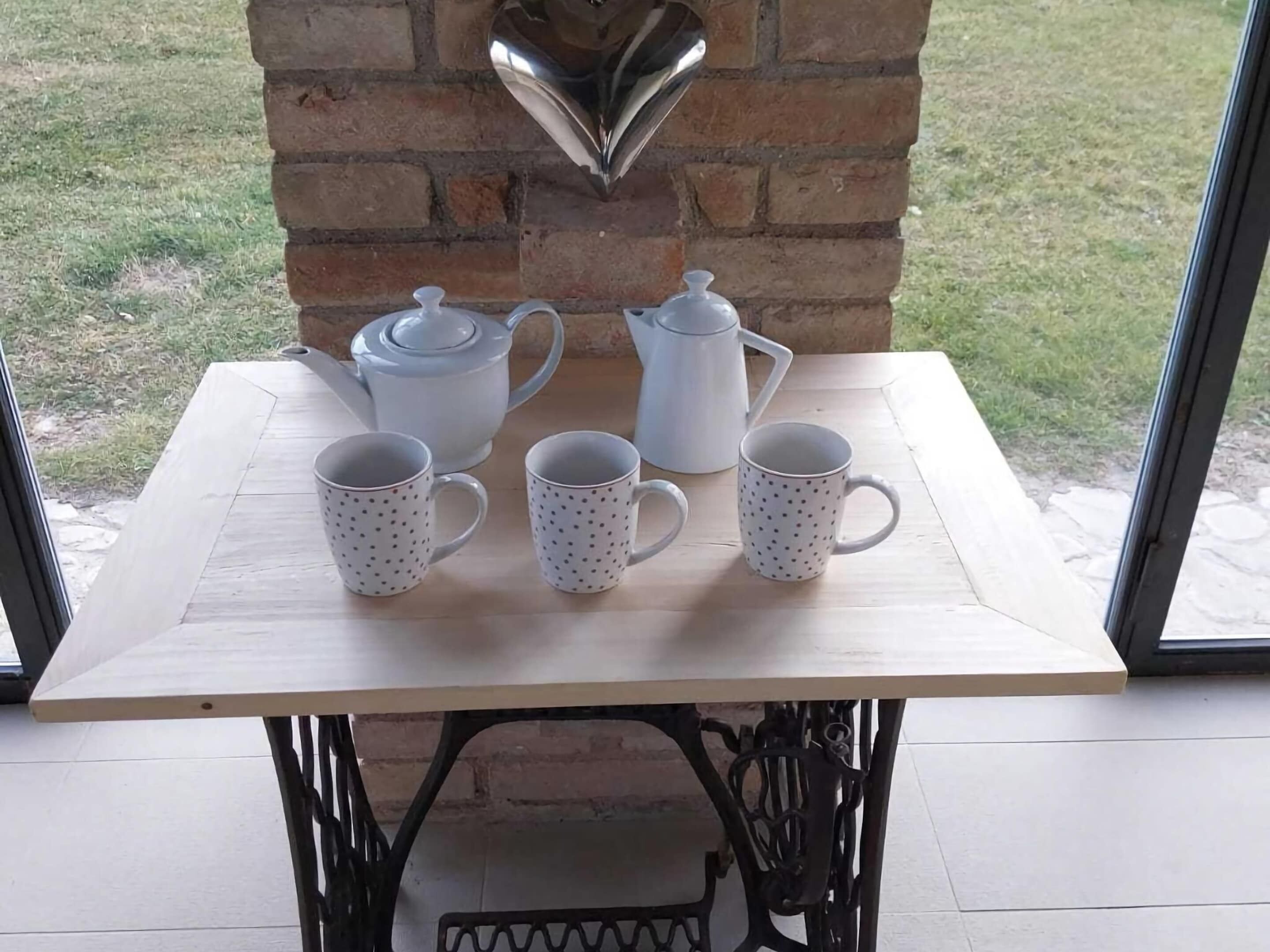 A table with a white teapot, two white cups, and a white sugar bowl on a wooden table with a brick wall behind it.