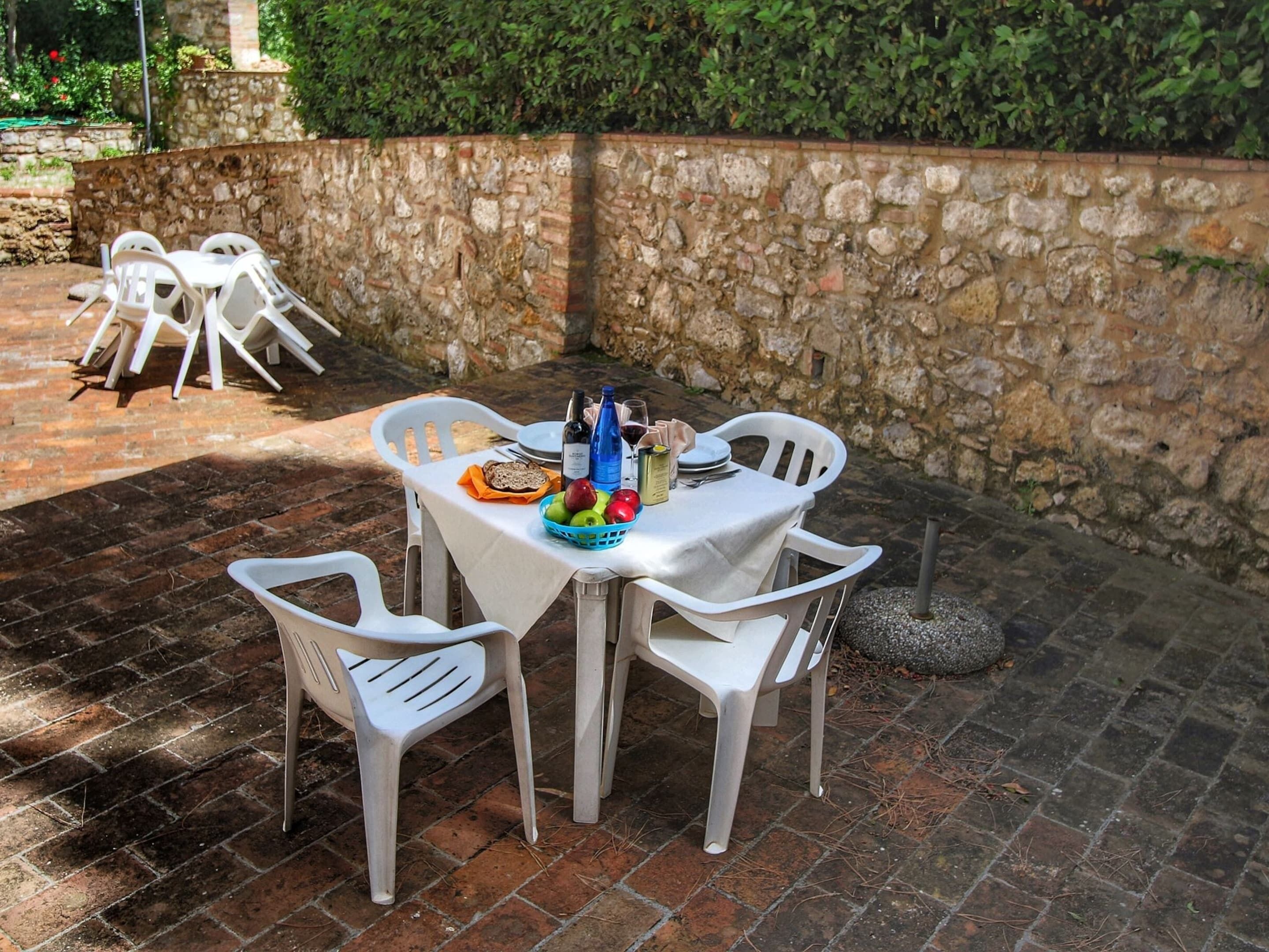 An outdoor dining area with a stone wall backdrop, white plastic chairs, and a table set with food and drinks.