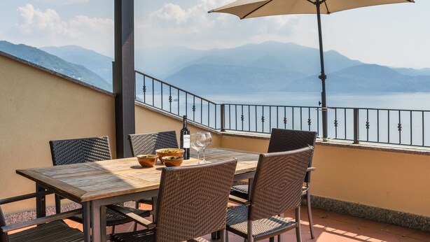Een terras met een tafel, stoelen, een parasol en uitzicht op bergen en een meer.