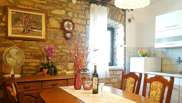 Zona pranzo con tavolo in legno, sedie, un vaso con rami, una bottiglia di vino e una sveglia appesa al muro.