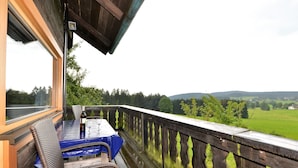 A balcony with a table and chairs, overlooking a green landscape with trees and hills. - Ferienhaus in Bayern mit Waldblick (Viechtach)