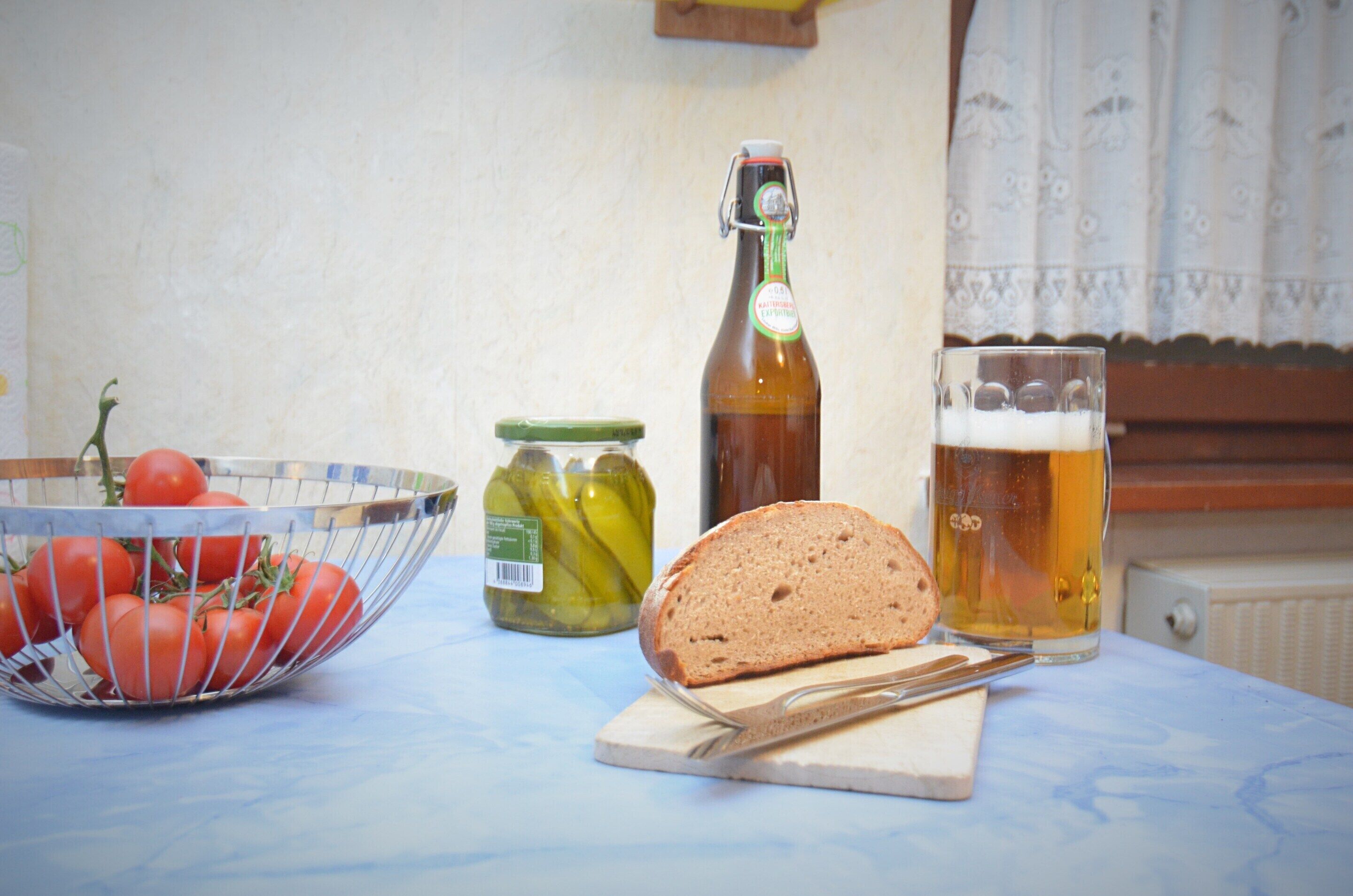 A table with a bowl of tomatoes, a loaf of bread, a bottle of beer, and a jar of pickles.