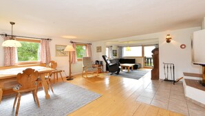 A spacious living area with a wooden dining table, chairs, a sofa, and a rocking chair. There is a large window with curtains, a lamp, and a wall-mounted TV. - Ferienhaus in Bayern mit Waldblick (Viechtach)