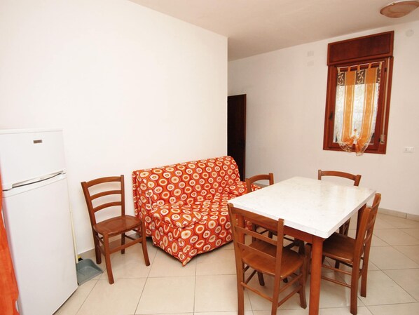 A small dining area with a white table and four wooden chairs, a patterned tablecloth, a white refrigerator, and a window with curtains.